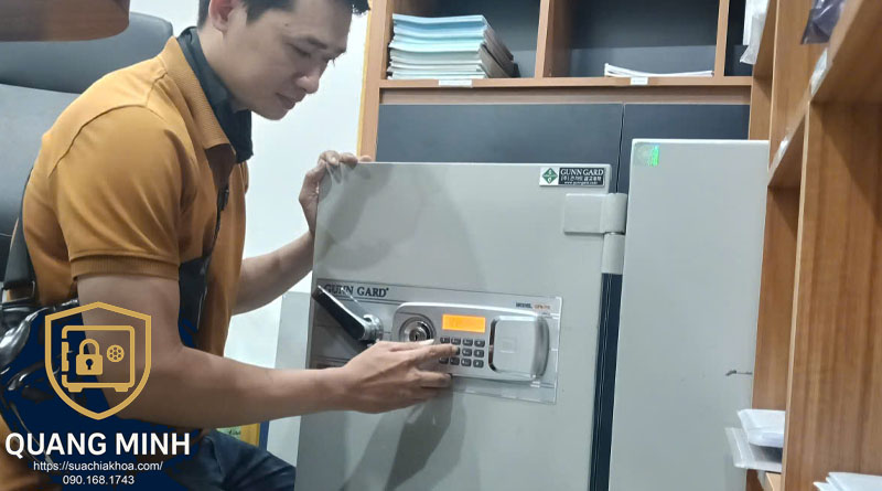 Người đàn ông áo vàng ngồi ghế văn phòng, một tay vịn nóc két Gunn Gard, tay kia bấm bàn phím