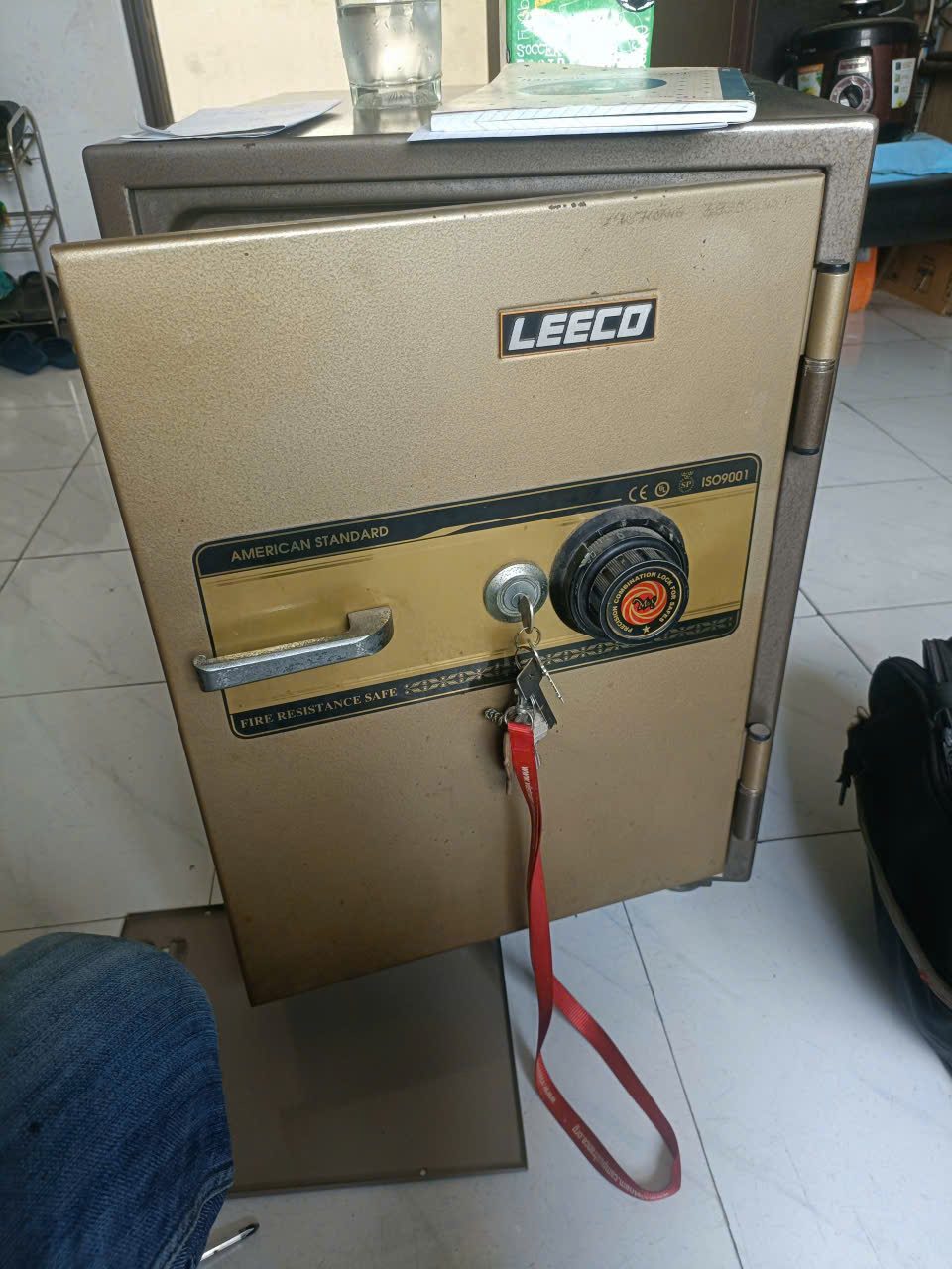 Két sắt cơ Leeco màu vàng đồng, chìa cắm sẵn trong ổ với chùm chìa và dây đeo đỏ treo lủng lẳng