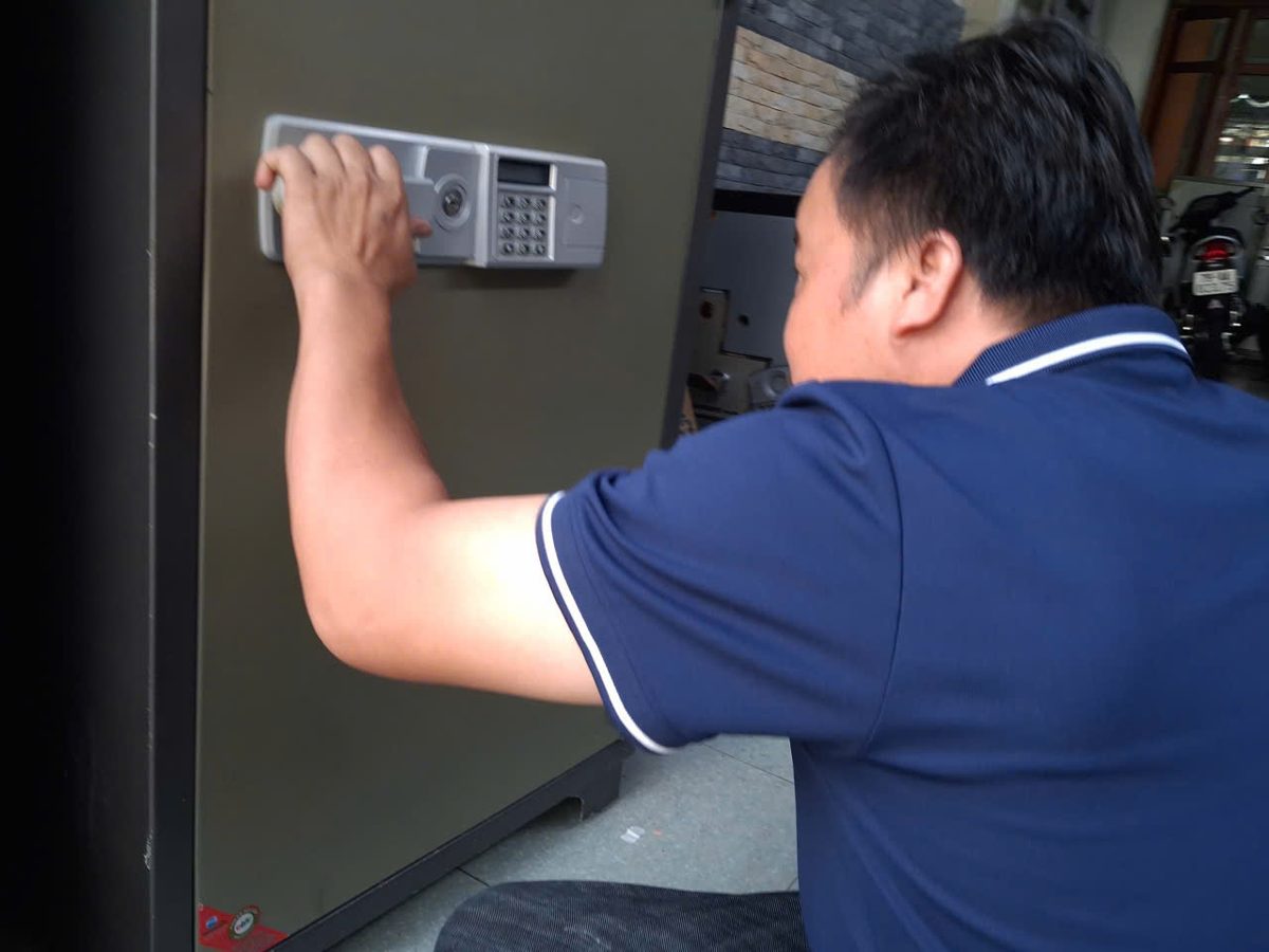 Thợ áo xanh đặt tay lên bảng khóa điện tử gắn trên cánh két sắt cỡ lớn để kiểm tra lỗi