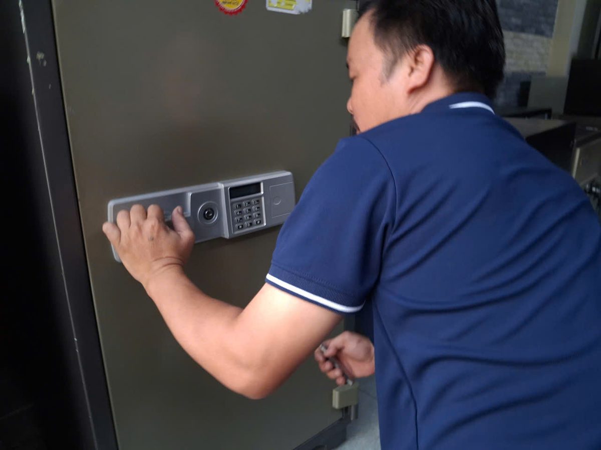 Thợ một tay bấm bàn phím két sắt điện tử lớn, tay kia cầm chìa xoay ổ khóa phụ phía dưới cánh
