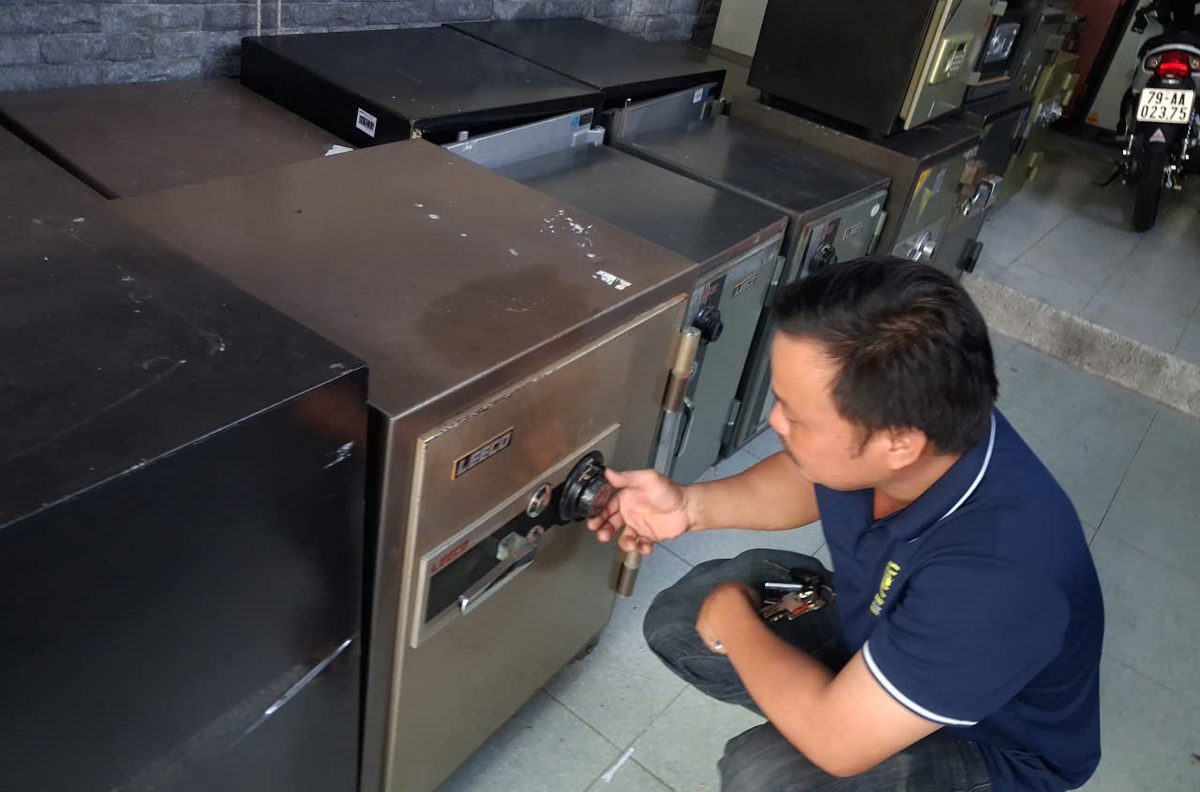 Thợ ngồi xổm cầm chùm chìa, tay kia xoay núm số két sắt cơ Leeco màu nâu