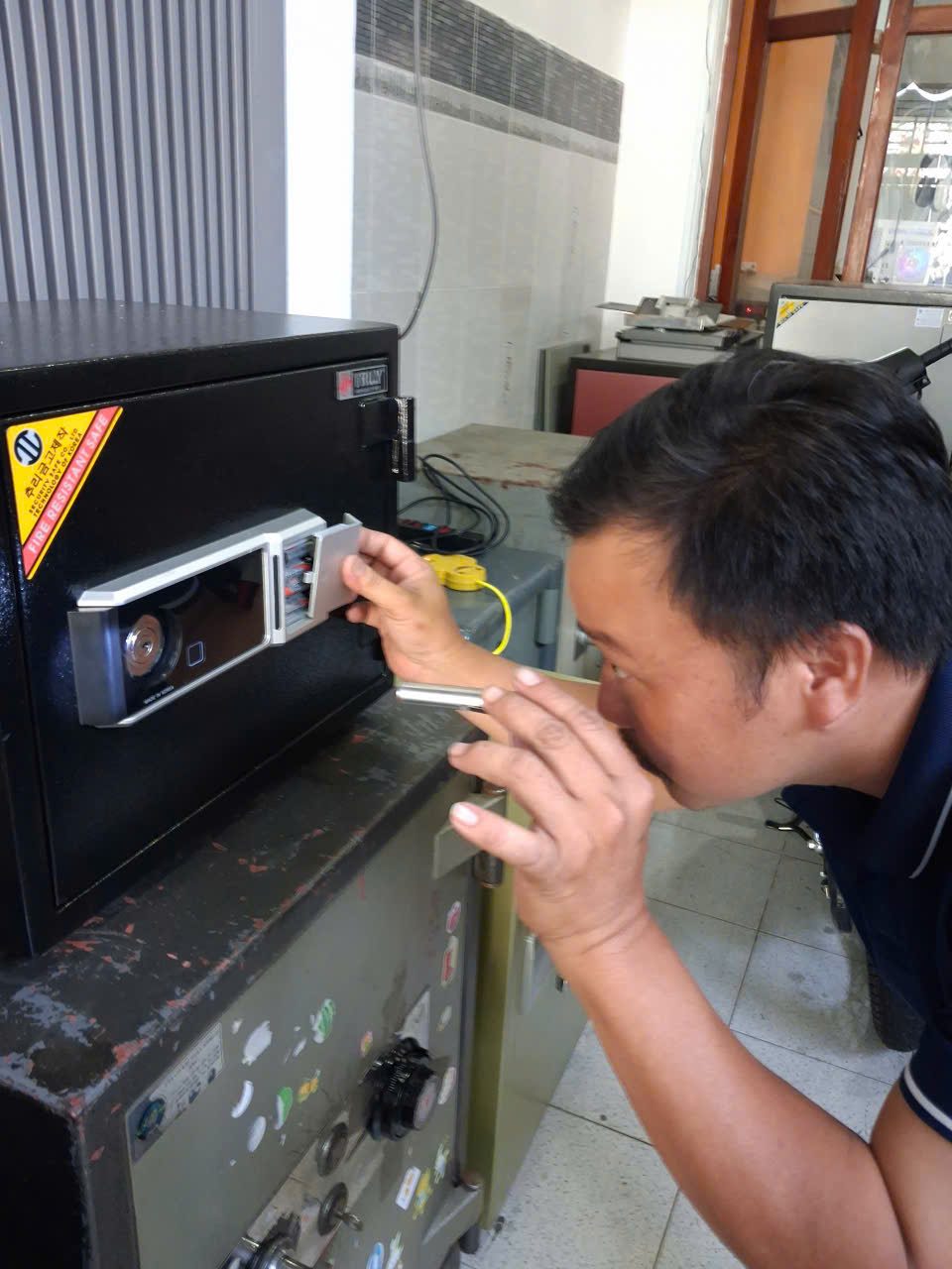 Thợ cúi sát, cầm đèn soi vào ngăn pin vừa kéo nắp trên bảng khóa két điện tử chống cháy màu đen