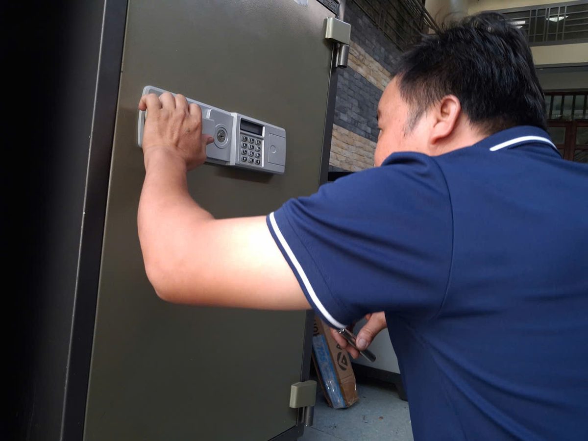 Thợ áo xanh đặt tay lên bảng khóa điện tử gắn trên cánh két sắt văn phòng cỡ lớn màu xám