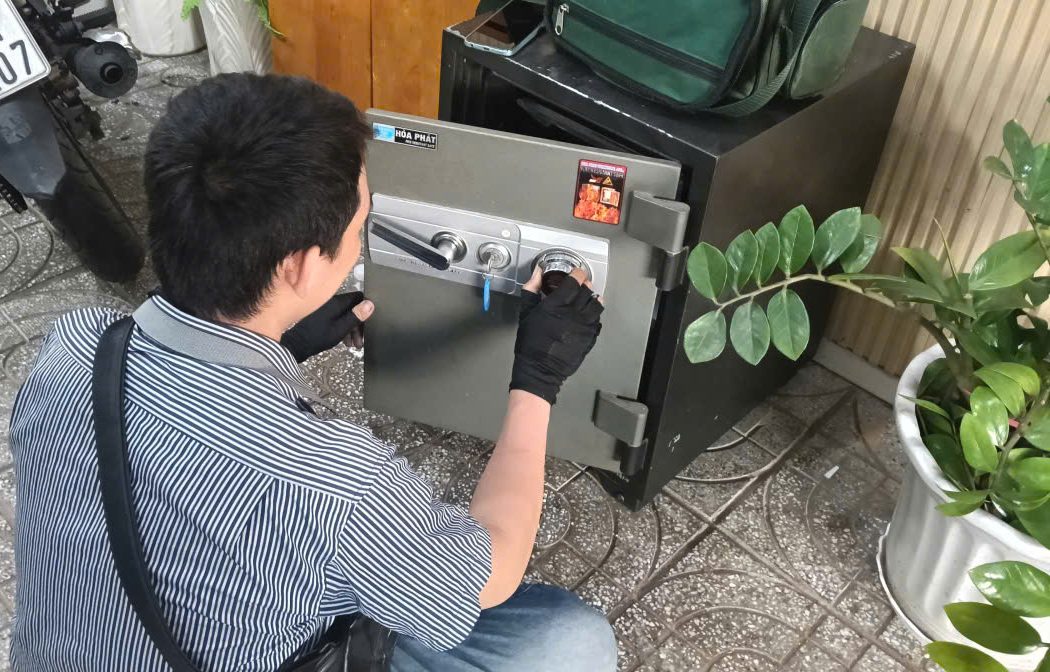 Thợ đeo găng đen ngồi xổm xoay núm số trên két sắt cơ Hòa Phát đã mở cửa, đặt ngoài hiên nhà