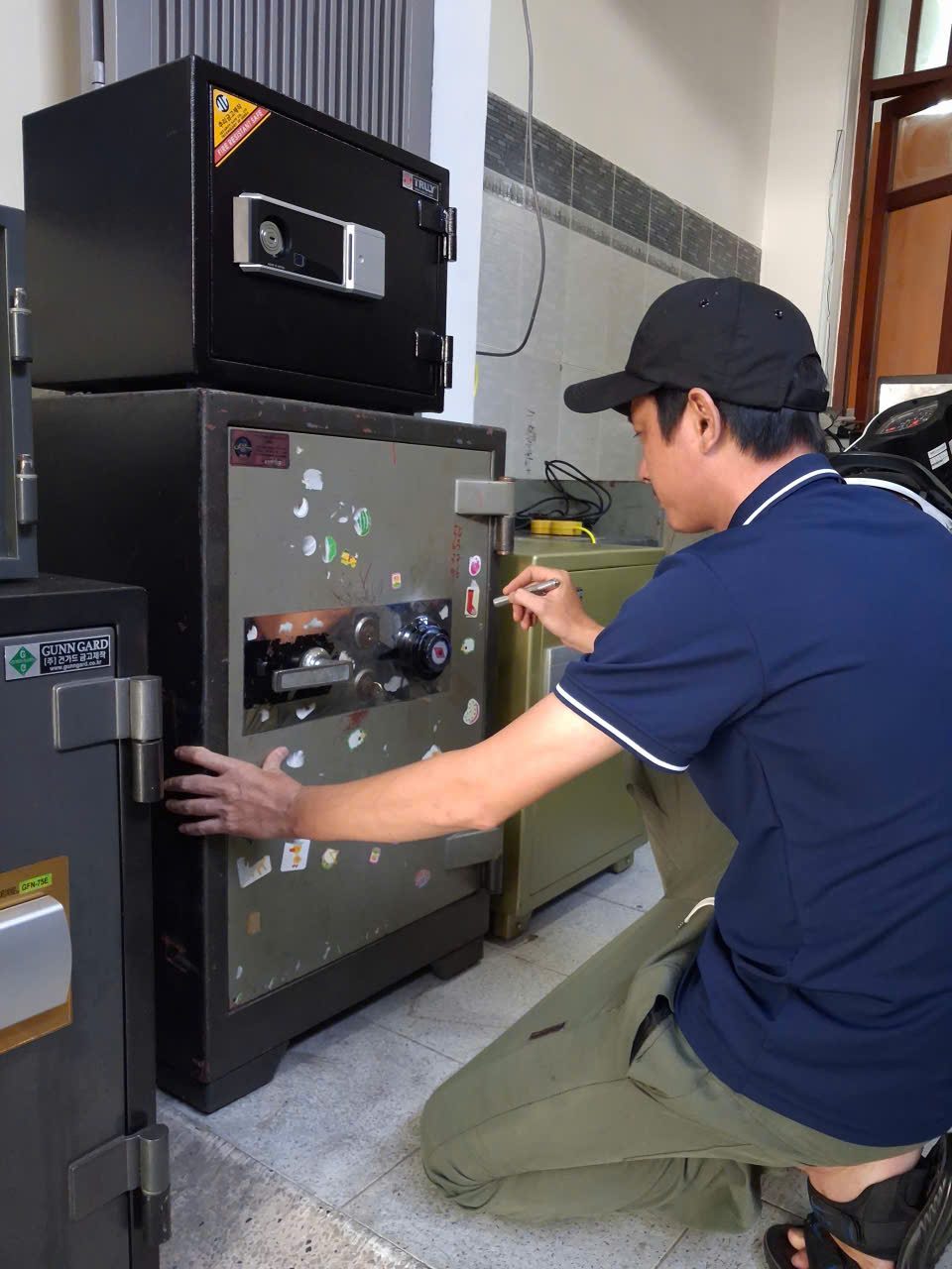Thợ đội mũ ngồi xổm, một tay đỡ cánh, tay kia cầm đèn pin soi vào núm xoay số của két cơ cũ