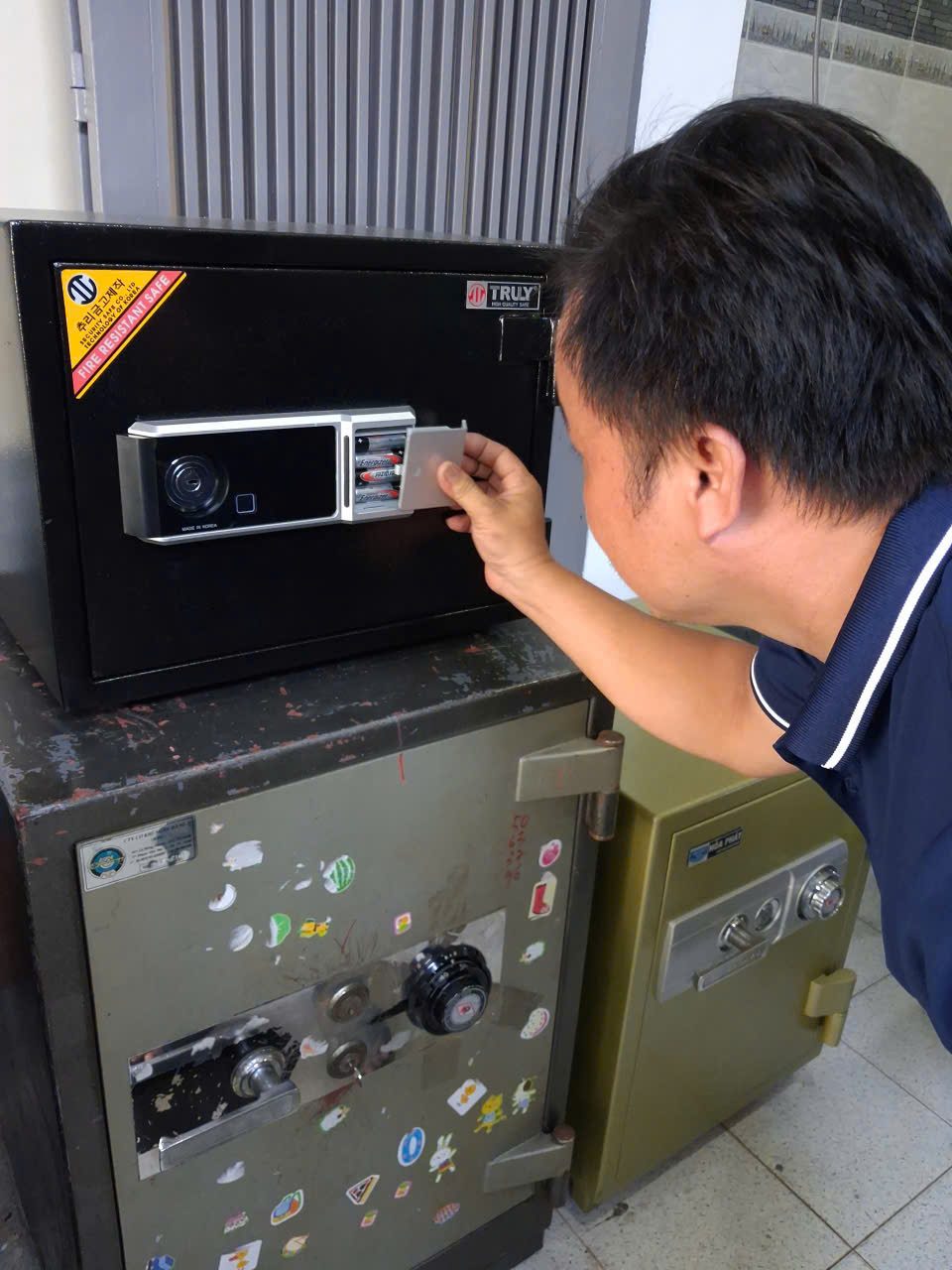 Thợ kéo nắp ngăn pin trên bảng khóa điện tử có ổ chìa và đầu đọc vân tay của két Truly màu đen