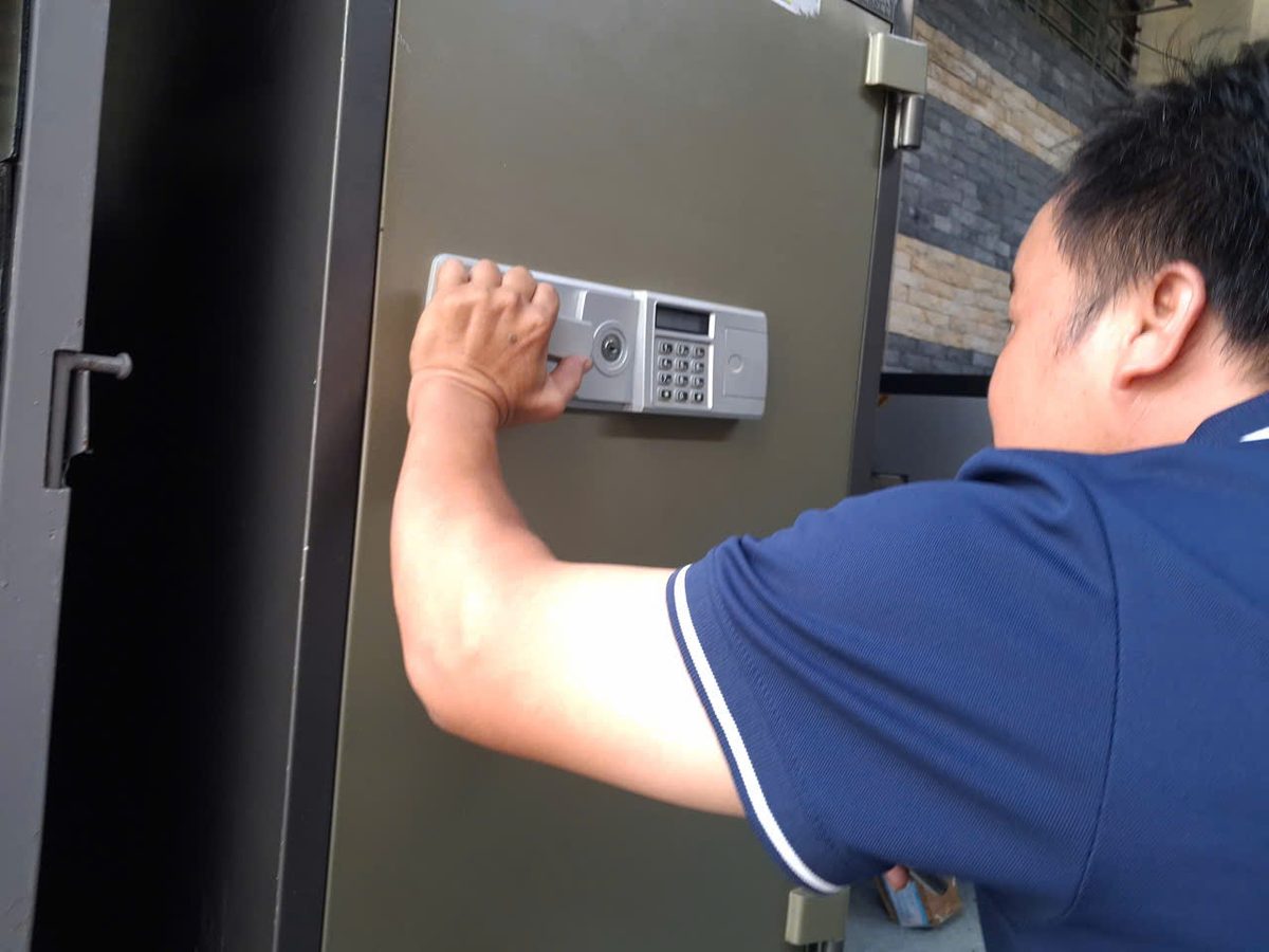 Thợ áo xanh đặt tay lên bảng khóa điện tử trên két cỡ lớn màu ghi, cánh két sát khung sắt bên trái