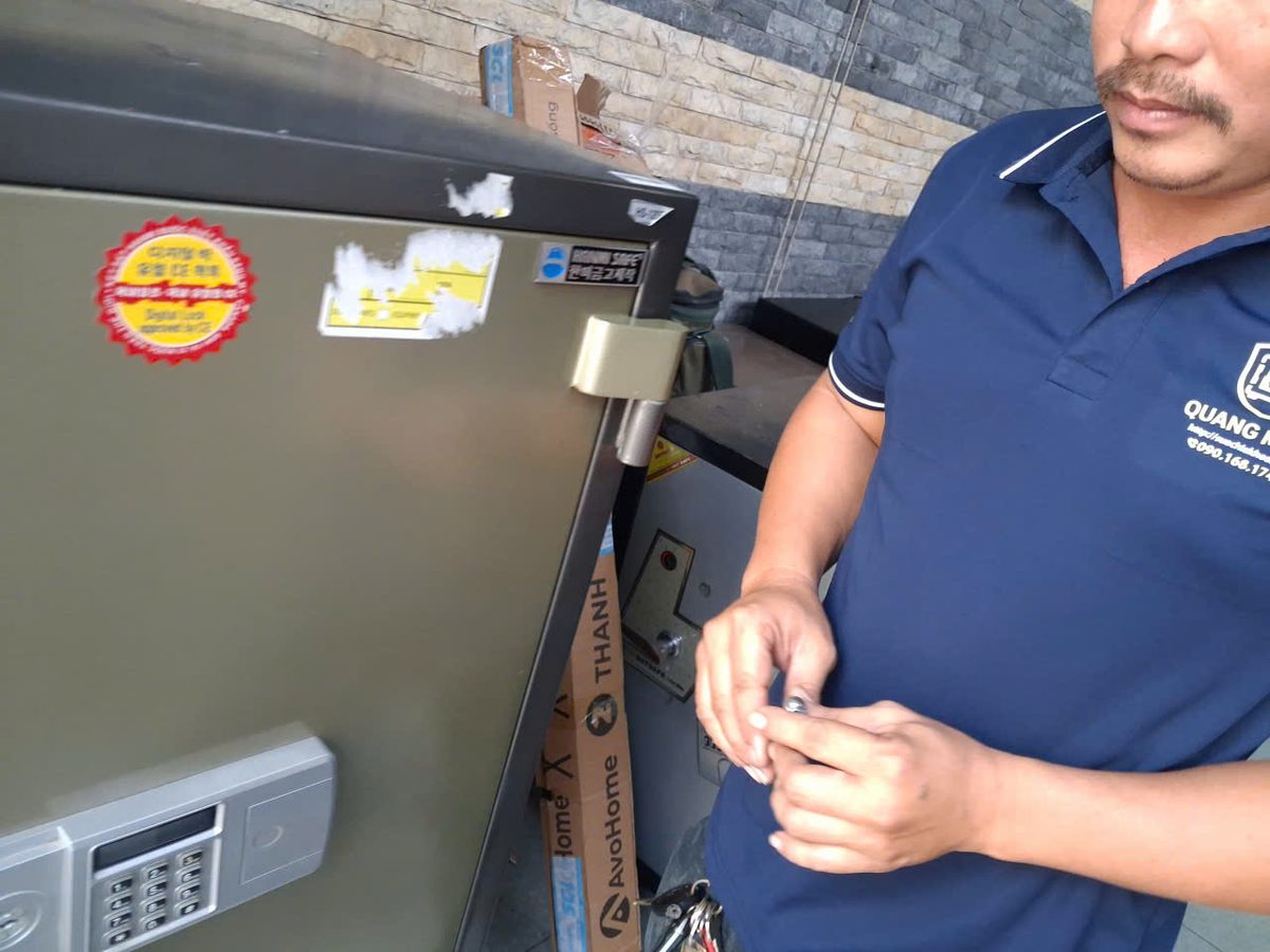Thợ áo Quang Minh cầm chìa khóa nhỏ đứng cạnh két sắt điện tử cỡ lớn màu xám